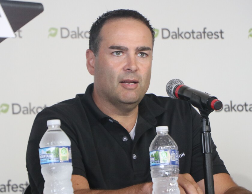 A man in a black polo shirt sits against a Dakotafest farm show backdrop, speaking into a microphone.