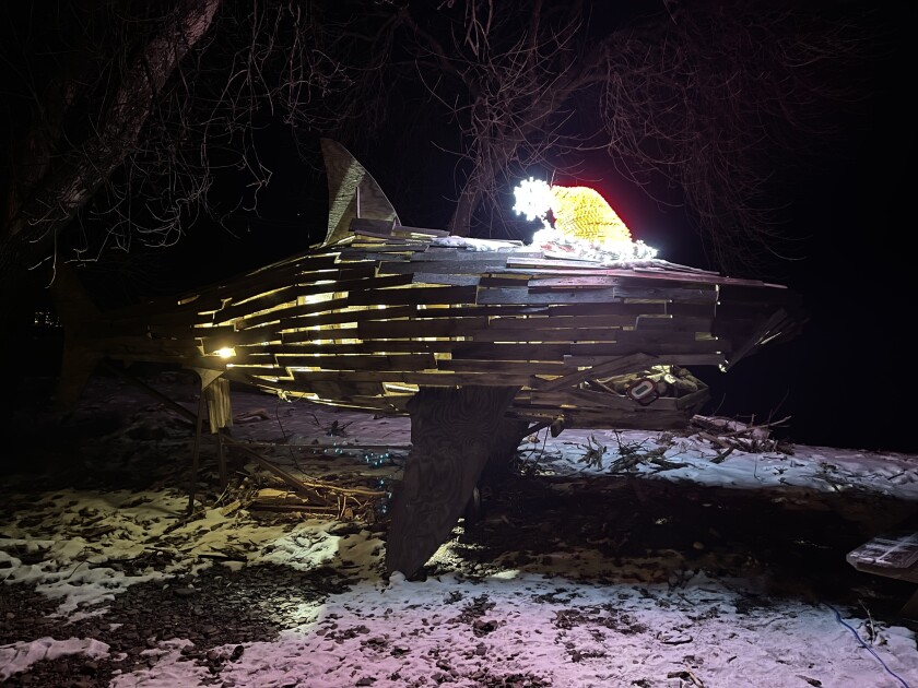 Large wooden shark sculpture is seen at night on a snow-dusted, rocky lake beach. Sculpture is lit from within, and wears an illuminated Santa hat.