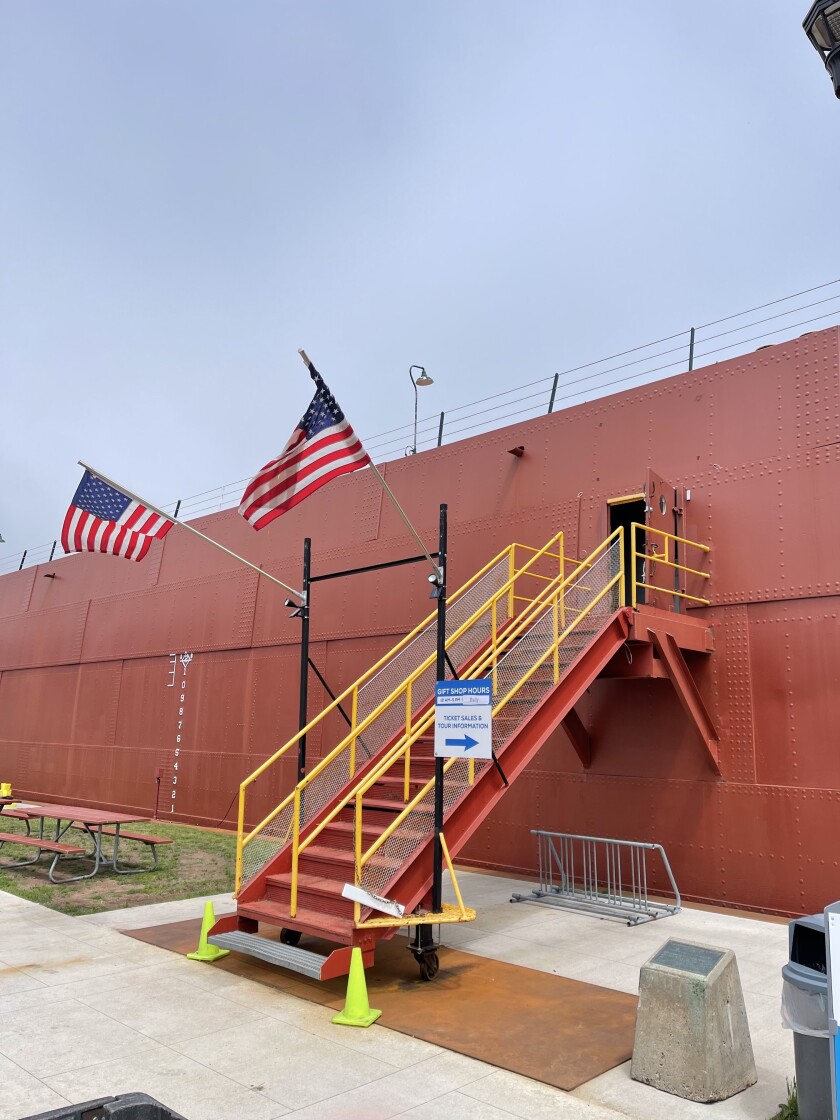 A ramp rises to the side of a large, brick red ship. Two American flags fly above the ramp; under the ramp, a bike rack is visible.