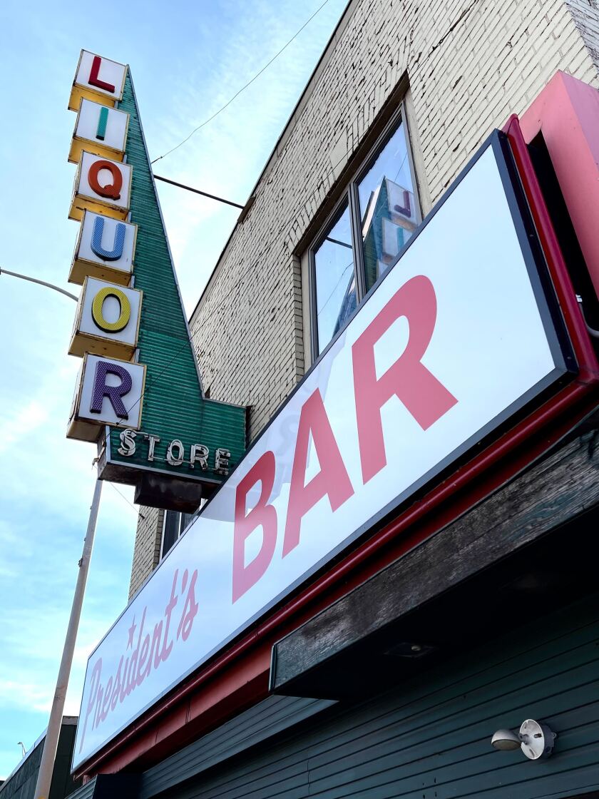 A horizontal sign reads "President's BAR" underneath a vertical sign reading, "LIQUOR STORE" in rainbow neon letters